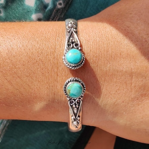 May include: A silver cuff bracelet featuring two round turquoise stones set at the open ends. The metalwork includes twisted rope detailing around each stone and decorative scrollwork patterns extending along the band toward the centre.