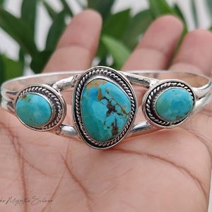 May include: A silver cuff bracelet featuring three turquoise stones with brown matrix patterns. The central stone is larger and oval-shaped, flanked by two smaller round stones. Each stone is set in a twisted rope-style silver bezel frame.