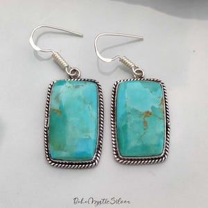 May include: A pair of rectangular drop earrings featuring polished turquoise stones with brown matrix veining. Each stone is set in a silver frame with a twisted rope border and attached to a silver fishhook wire. The text Boho Mystic Silver is visible.