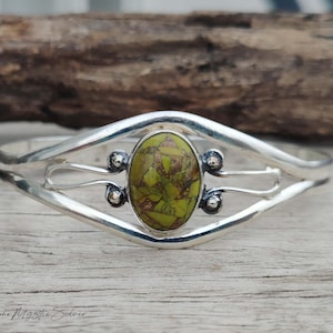 May include: A silver cuff bracelet featuring a central oval-shaped stone with a mosaic pattern of lime green and brown fragments. The metal band has a split design with decorative silver spheres on either side of the stone setting.