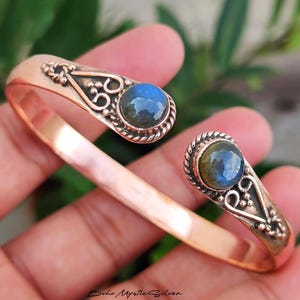 May include: A copper cuff bracelet featuring an open design with two rounded ends. Each end is set with a round, iridescent blue labradorite gemstone, surrounded by decorative twisted metal wire and small metal bead accents.