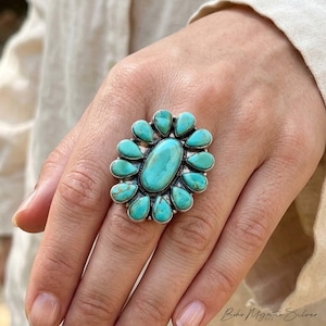 May include: A silver ring featuring a large central oval turquoise stone surrounded by twelve smaller, teardrop-shaped turquoise stones arranged in a floral pattern. The metal setting has a polished finish with visible oxidation in the crevices.