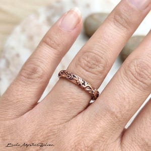 Peut inclure: Une bague en métal de couleur cuivre avec un motif floral et végétal en relief qui fait tout le tour de l'anneau. La surface de la bague présente une finition oxydée et sombre dans les creux du motif botanique complexe.
