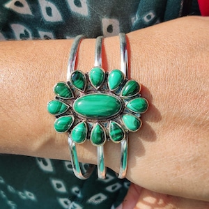 May include: A silver cuff bracelet featuring a large central oval malachite stone surrounded by twelve smaller teardrop-shaped malachite stones. The metal setting has a textured, oxidized finish, and the band consists of three parallel silver wires.