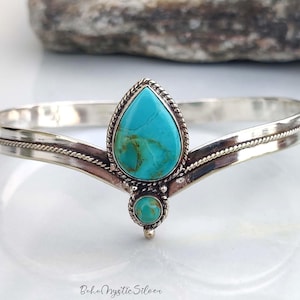 May include: A silver cuff bracelet featuring a V-shaped band with a twisted rope texture. The centre is adorned with a large pear-shaped turquoise stone and a smaller round turquoise stone, both set in silver bezels with decorative rope detailing.
