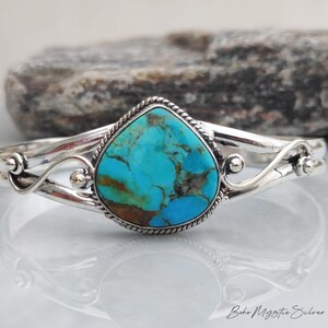 May include: A silver cuff bracelet featuring a central pear-shaped turquoise stone with brown matrix veining. The stone is framed by a twisted rope border, and the band displays decorative silver scrollwork and small spheres on each side.