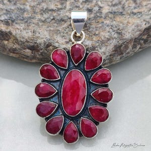 May include: A silver pendant featuring a large, central oval-shaped red gemstone surrounded by twelve smaller, teardrop-shaped red gemstones. The metal setting has an oxidised finish, and a silver bail is attached to the top of the piece.