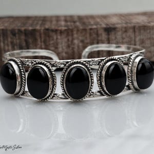 May include: A silver cuff bracelet featuring five oval-shaped black stones set in rope-textured bezels. The band is constructed from two parallel silver wires with a floral pattern and an open back design. The text Boho Mystic Silver is visible.