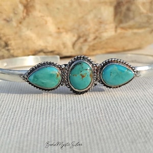 Könnte beinhalten: Ein silberner Armreif mit drei Türkissteinen. Der mittlere Stein ist oval und wird von zwei birnenförmigen Steinen flankiert. Jeder Stein ist in eine gedrehte Silberkordel eingefasst. Der Text "Boho Mystic Silver" steht unten im Bild.
