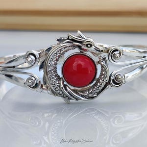 May include: A silver cuff bracelet featuring a central circular red stone framed by a detailed dragon motif with textured scales and swirling metal accents. The metal has a polished finish, and the text Boho Mystic Silver is visible at the bottom.