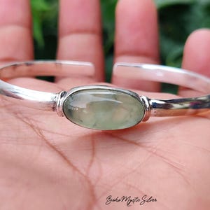 May include: A silver cuff bracelet featuring a smooth, polished finish and an open-ended design. The center of the bracelet is set with a single, oval-shaped, translucent pale green gemstone with subtle internal inclusions and a polished surface.