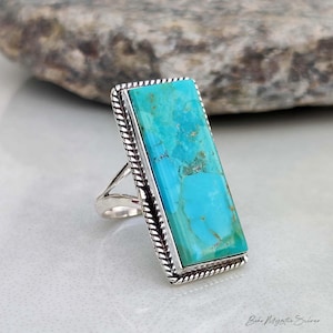 May include: A rectangular turquoise stone set in a silver ring with a twisted rope border design. The stone features a vibrant blue-green color with natural brown matrix veining. The silver band has a split shank design at the base.