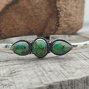 May include: A silver cuff bracelet featuring three green turquoise stones with brown matrix patterns. The centre stone is oval, flanked by two pear-shaped stones, each set in a twisted silver rope border. The text Boho Mystic Silver is visible.