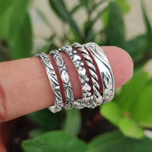 May include: A set of five silver rings worn on a finger. The rings feature distinct textures including a leaf pattern, a geometric diamond design, a row of small hearts, a twisted rope band, and a smooth band with an engraved floral motif.