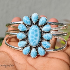 May include: A silver cuff bracelet featuring a large oval central larimar stone surrounded by twelve smaller pear-shaped larimar stones in a floral arrangement. The metal setting has a polished finish, and the text Boho Mystic Silver is visible.