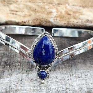 May include: A silver cuff bracelet featuring a V-shaped design with a central teardrop-shaped lapis lazuli stone and a smaller round lapis lazuli stone below it. The silver band has a polished finish with twisted wire detailing around the stones.
