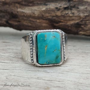May include: A silver ring featuring a rectangular turquoise stone with brown matrix veining. The stone is set in a smooth bezel flanked by two vertical rows of small silver beads. The band is wide and polished, with the text Boho Mystic Silver.