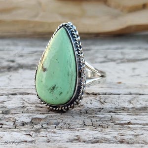May include: A teardrop-shaped light green stone ring set in a polished silver band. The stone features subtle dark inclusions and a smooth, glossy surface. The setting is framed by a decorative twisted rope-style silver border detail.
