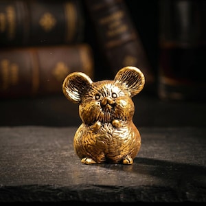 May include: A small, metallic gold-colored figurine of a mouse with large, rounded ears, a textured coat, and a rounded body. The mouse is depicted in a standing position with its front paws tucked near its chest, featuring detailed fur markings.