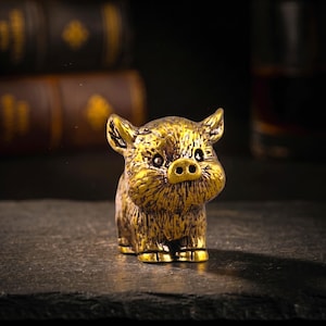Peut inclure: Une petite figurine en métal doré représentant un porcelet avec une finition texturée et vieillie. Le porcelet a de grandes oreilles pointues, un museau arrondi avec deux narines et des détails sombres en creux soulignant sa texture.