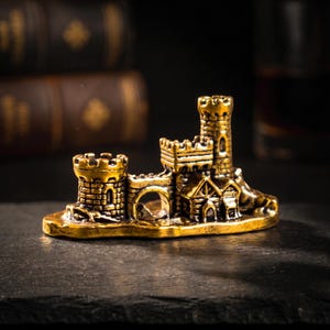 May include: A miniature gold-colored metal castle figurine featuring two distinct towers, a central arched gateway, and detailed brickwork textures. The structure is mounted on a flat, irregular base with a metallic finish and visible relief.