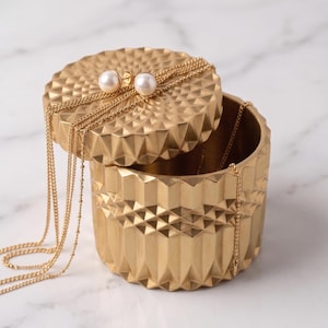 May include: A cylindrical gold-toned container with a textured geometric pattern and a matching lid. A pair of white pearl stud earrings and several delicate gold chains are draped over the lid and inside the open container.