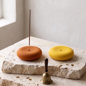 May include: Two smooth, rounded incense holders in matte terracotta orange and bright yellow, each featuring a central hole. A thin brown incense stick stands upright in the orange holder. A small brass hand bell with a dark wooden handle sits nearby.