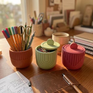 May include: Three rounded containers with vertical ribbed textures in terracotta, sage green, and dusty rose. The terracotta container holds various colored pens and gold scissors. The sage green and dusty rose containers have matching lids.