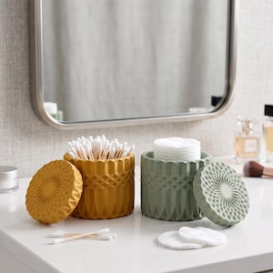 May include: Two cylindrical containers with geometric faceted patterns on the exterior. One container is mustard yellow and filled with cotton swabs, while the other is sage green and holds a stack of round white cotton pads, each with a matching lid.
