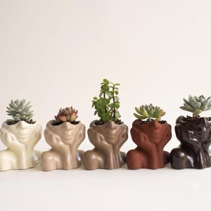 May include: Five ceramic planters shaped like human heads resting on hands, arranged in a row. Each planter features a different skin tone, ranging from cream to dark brown, and contains a small green succulent plant growing from the top.