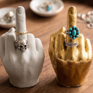 May include: Two hand-shaped ring holders, one matte white and one metallic gold, each displaying a middle finger gesture. The white holder features a thin gold band and a silver skull ring, while the gold holder displays a turquoise cluster ring.