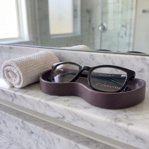 May include: A pair of black-framed eyeglasses resting inside a smooth, dark purple, oval-shaped tray. Next to the tray sits a small, white, waffle-textured rolled towel. The items are positioned on a white marble surface with gray veining.
