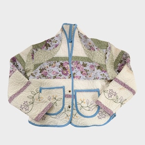 May include: A quilted jacket featuring a cream-colored base with patchwork panels of purple floral patterns and green textured fabric. The garment includes two front pockets with embroidered floral vines, light blue trim, and metal snap closures.