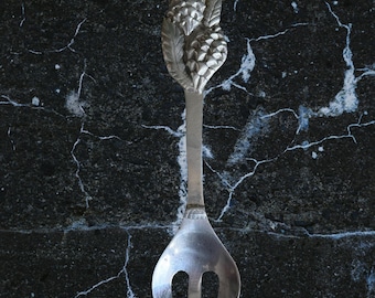 May include: A silver-toned metal serving fork featuring a handle decorated with a detailed relief of two pinecones and foliage. The utensil has a smooth, polished finish on the three-pronged head and a textured, matte handle design.