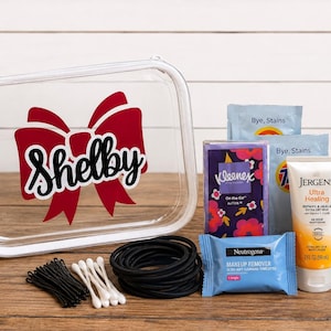 May include: A clear rectangular plastic case with a red bow graphic and the name Shelby printed in black. The set includes black bobby pins, white cotton swabs, black hair ties, Kleenex tissues, Tide stain remover, Neutrogena wipes, Jergens lotion, Marel spray, and Rave hairspray.