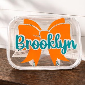May include: A clear rectangular plastic pouch featuring a large orange bow graphic with the name Brooklyn printed in a teal script font across the center. The pouch is transparent, allowing the bow design to be clearly visible through the material.