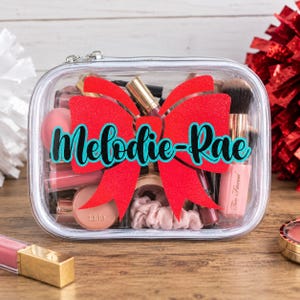 May include: A clear rectangular cosmetic bag with a zipper closure, featuring a large red glitter bow graphic and the name Melodie-Rae printed in teal script. The bag is filled with various makeup items including brushes and tubes.