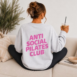 May include: A light gray crewneck sweatshirt featuring the text ANTI SOCIAL PILATES CLUB printed in bold pink letters on the back. The garment has long sleeves and a relaxed fit, shown from a rear perspective while worn with black leggings.