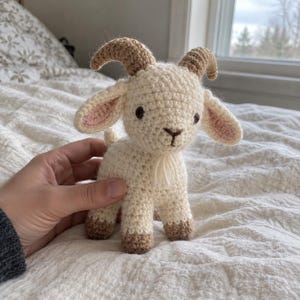 May include: A small, handmade crochet goat toy featuring cream-coloured yarn for the body, light brown yarn for the curved horns and hooves, and soft pink yarn detailing inside the ears. The goat has dark brown embroidered eyes and a small beard.