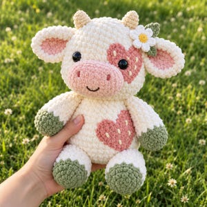 Pode incluir: Uma pelúcia de vaca em crochê na cor creme com manchas rosas em formato de coração no rosto e no peito, cascos verdes e uma flor de margarida branca com folhas verdes na cabeça. O brinquedo tem olhos pretos e um sorriso bordado.