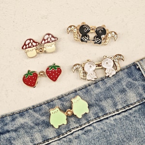 Peut inclure: Cinq ensembles de broches en métal doré reliées par une petite chaîne. Les motifs incluent deux champignons rouges et blancs, deux fraises rouges avec des feuilles vertes, deux écureuils noirs, deux écureuils blancs avec strass et deux grenouilles vert clair.