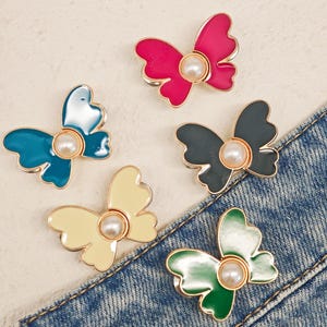 Peut inclure: Cinq broches en forme de papillon avec des bords en métal doré et une perle blanche centrale. Les broches présentent des finitions en émail aux couleurs vives, notamment rose fuchsia, bleu sarcelle, gris foncé, crème et un dégradé de vert.