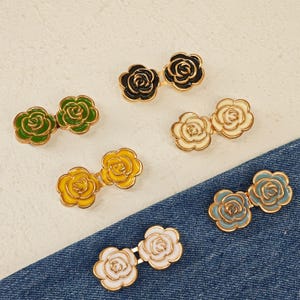 Peut inclure: Une collection de six pinces à cheveux en forme de double fleur, chacune composée de deux breloques roses avec des bords en métal doré. L'ensemble comprend des couleurs noir, vert, crème, jaune, bleu clair et blanc sur une surface texturée.
