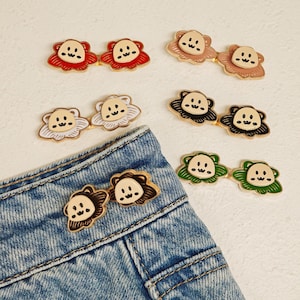 Peut inclure: Six paires de broches en émail métallique en forme de visages de dessin animé avec des cheveux ondulés, aux couleurs rouge, beige, blanc, noir, marron et vert. Une paire est fixée à la ceinture d'un jean bleu, cinq paires sont au-dessus.