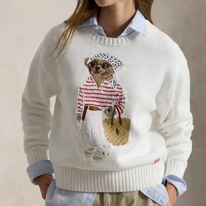 Könnte beinhalten: Ein weißer Langarmpullover mit Rundhalsausschnitt und einer gestickten Bärengrafik. Der Bär trägt ein weiß-blau gepunktetes Kopftuch, eine Sonnenbrille, ein rot-weiß gestreiftes Hemd, eine weiße Hose und eine beige geflochtene Tasche.