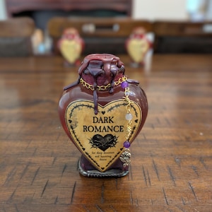 May include: A heart-shaped glass bottle with dark red liquid, topped with dark wax drips. The label reads DARK ROMANCE for deep devotion and haunting desire, framed by barbed wire. A gold chain with purple beads and a rose charm hangs from the neck.
