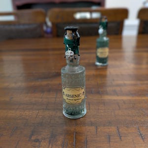 May include: A clear glass bottle with a cork stopper sealed in black wax, wrapped in green twine with a silver skull charm. The label reads ARSENIC all-natural ingredients with skull icons. A second similar bottle is visible in the background.