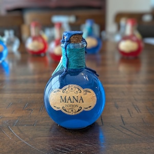 May include: A round glass bottle filled with blue liquid, featuring a cork stopper sealed with dark blue wax. The neck is wrapped in teal twine, and a beige label on the front displays the text MANA POTION in a decorative black font.