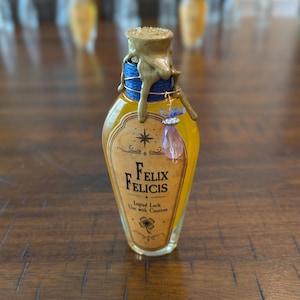 May include: A glass bottle filled with yellow liquid, featuring a label that reads Felix Felicis, Liquid Luck, Use with Caution. The bottle neck is wrapped in blue cord and topped with gold wax, with a small purple crystal charm attached by wire.