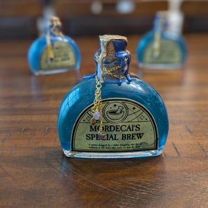 May include: A glass bottle filled with blue liquid, featuring a cork stopper sealed with gold and blue wax. A gold chain with small beads hangs from the neck. The label reads: MORDECAI'S SPECIAL BREW, A potion designed by a shifty changeling.